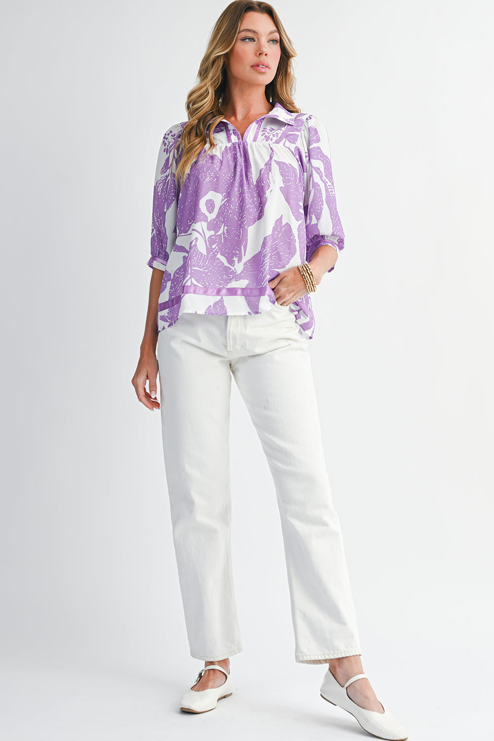 Woman wearing a purple patterned blouse and white pants on a plain background