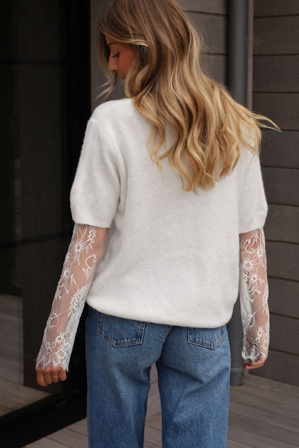 Person wearing a white sweater with lace sleeves and blue jeans, standing against a wooden wall.