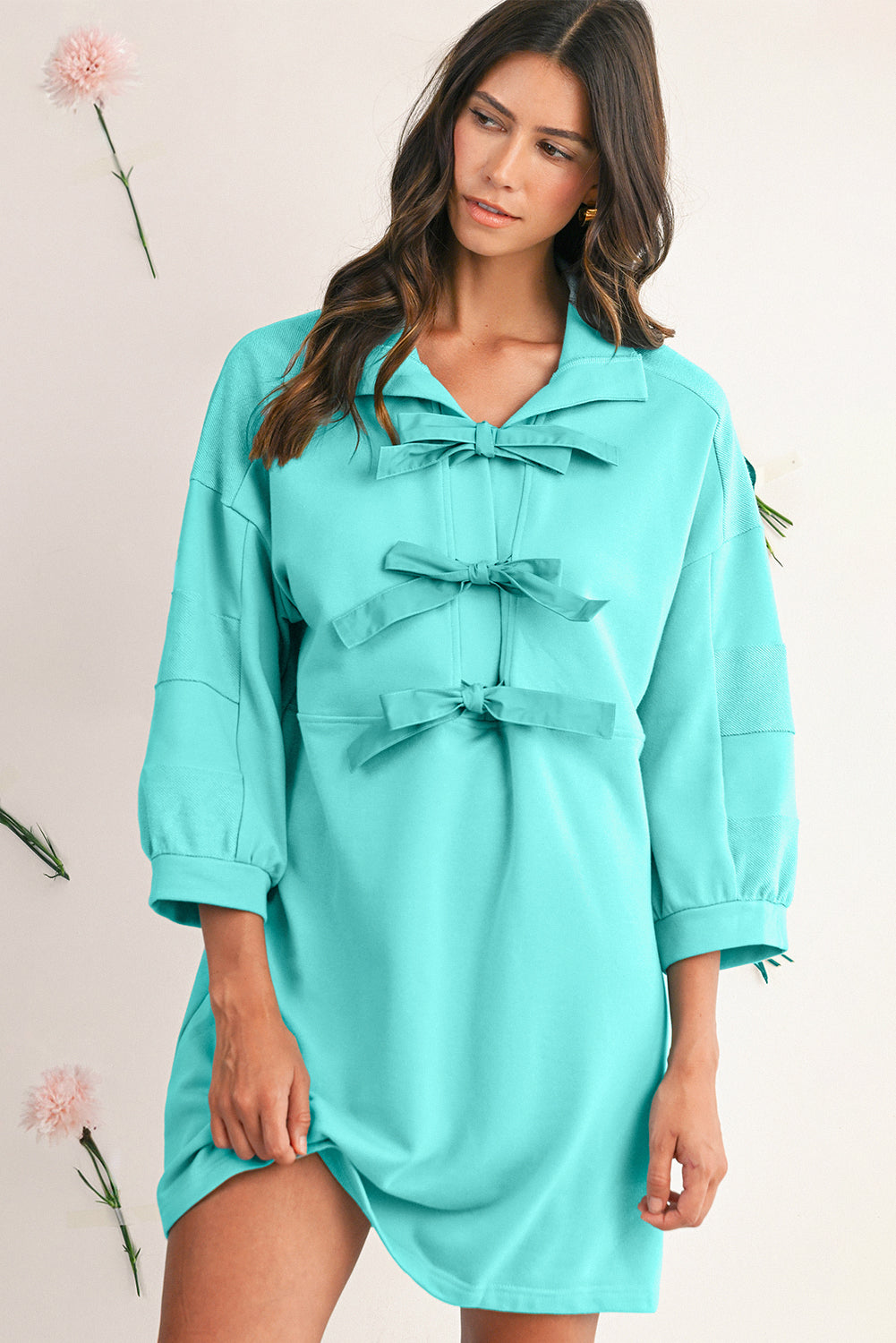 Woman wearing a turquoise dress with long sleeves and bow details on a plain background