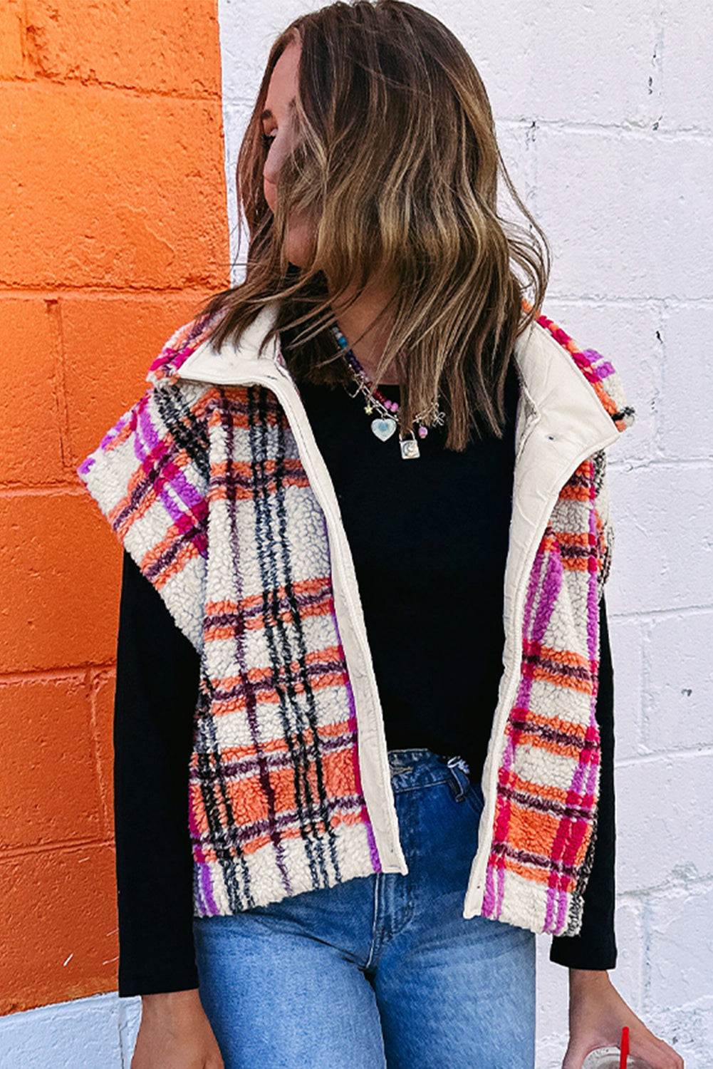 Woman wearing a plaid vest over a black top and blue jeans against an orange and white wall.