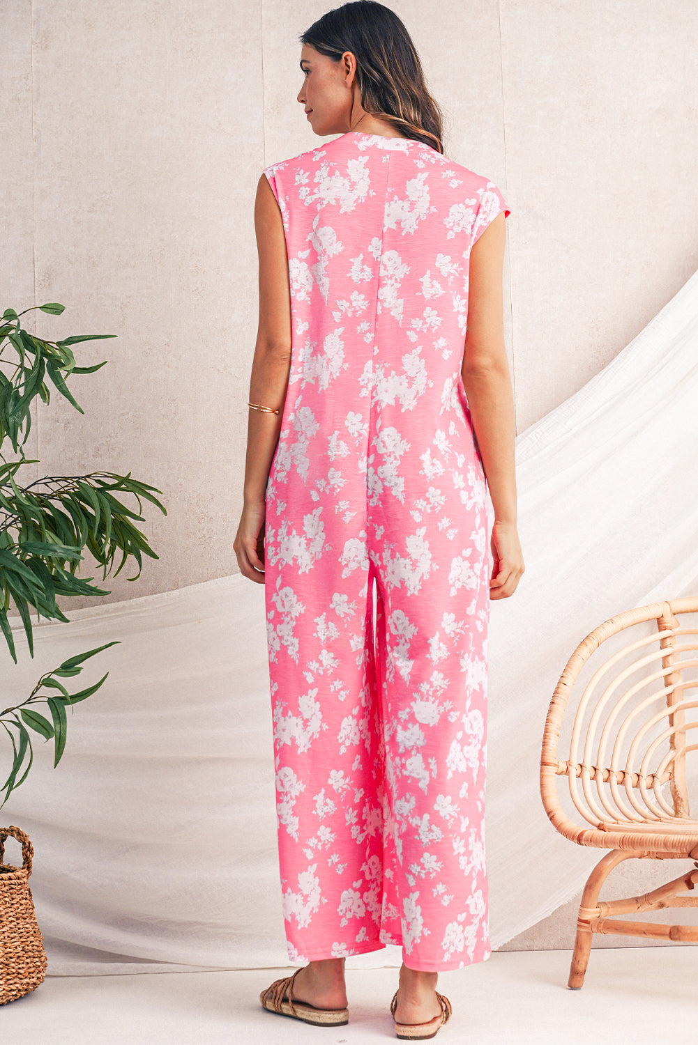 Woman wearing a pink floral jumpsuit standing in a minimalistic room with a plant and chair.