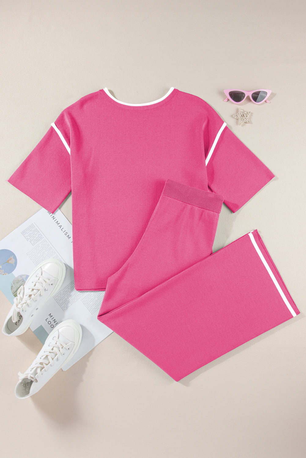 Pink pajama set with white accents on a beige surface, accompanied by white sneakers and sunglasses.