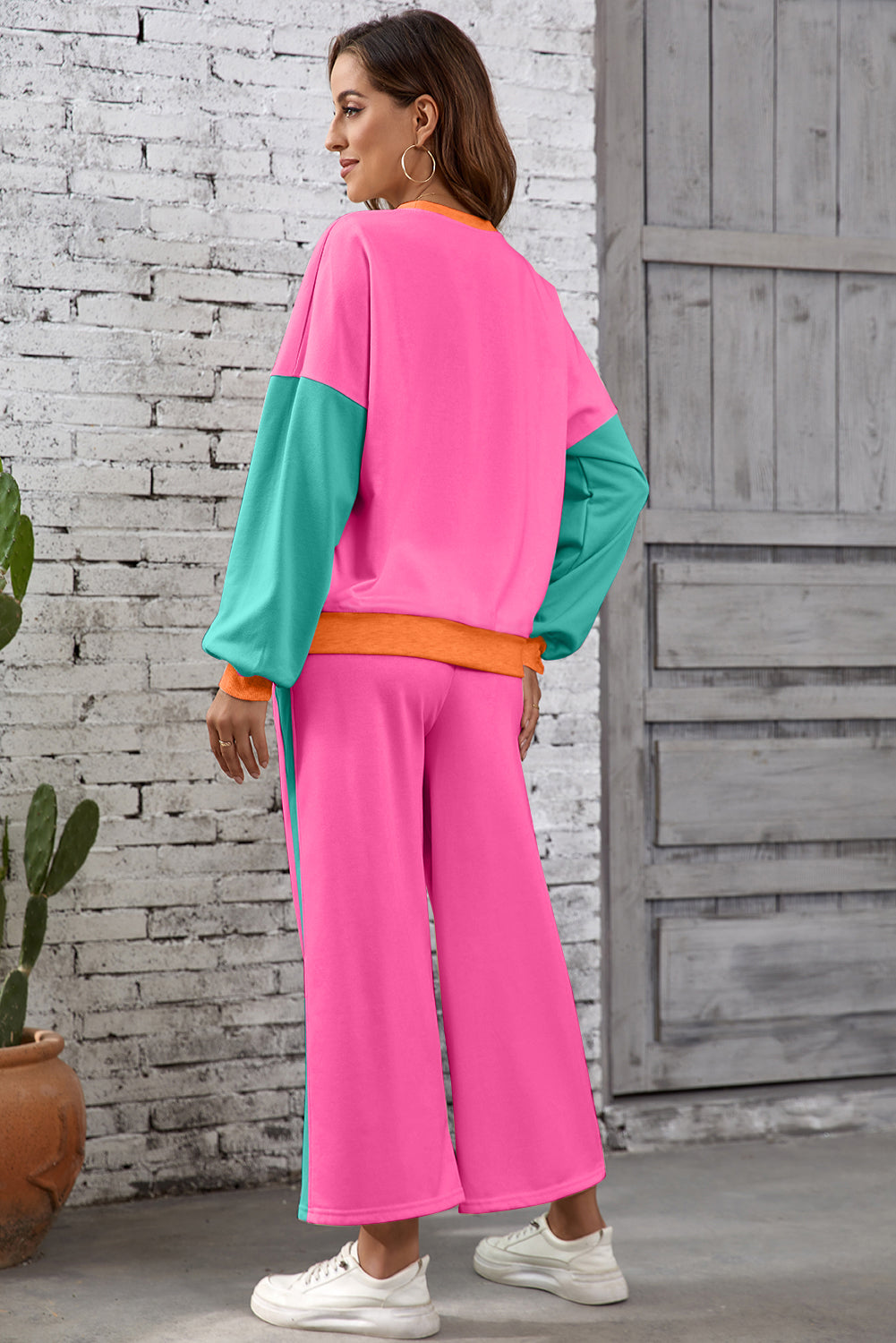 Woman wearing a colorful outfit with pink and teal top and pink pants against a brick wall.