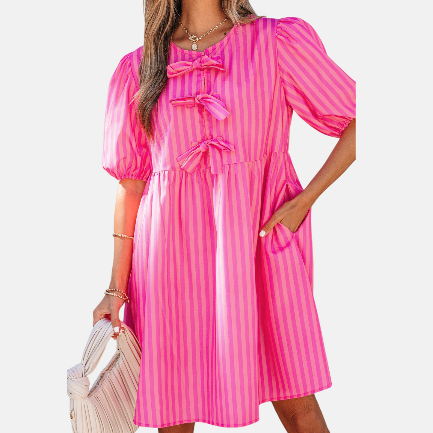 A model posing in a vibrant pink striped mini dress featuring puff sleeves, three decorative front bows, and functional side pockets, styled with a pleated handbag.