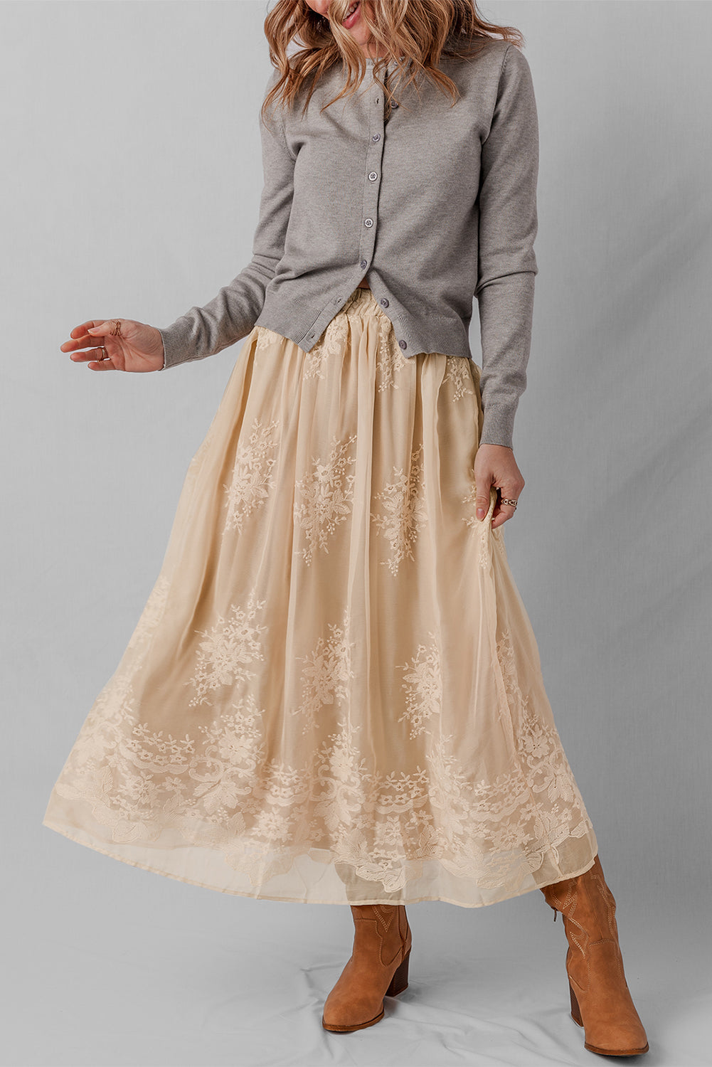 Woman wearing a gray shirt and beige lace skirt with brown boots on a gray background