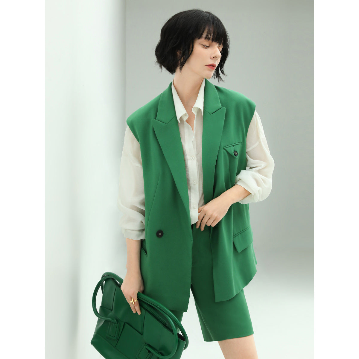 Woman wearing a green vest and shorts set with a white shirt, holding a green bag against a light gray background.