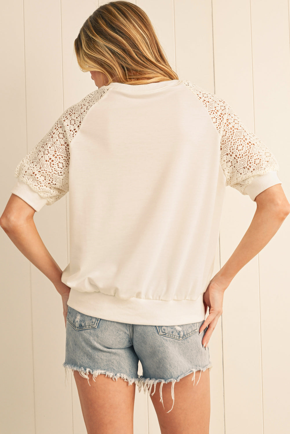 Woman wearing a white lace-trimmed top and denim shorts against a light wooden panel background