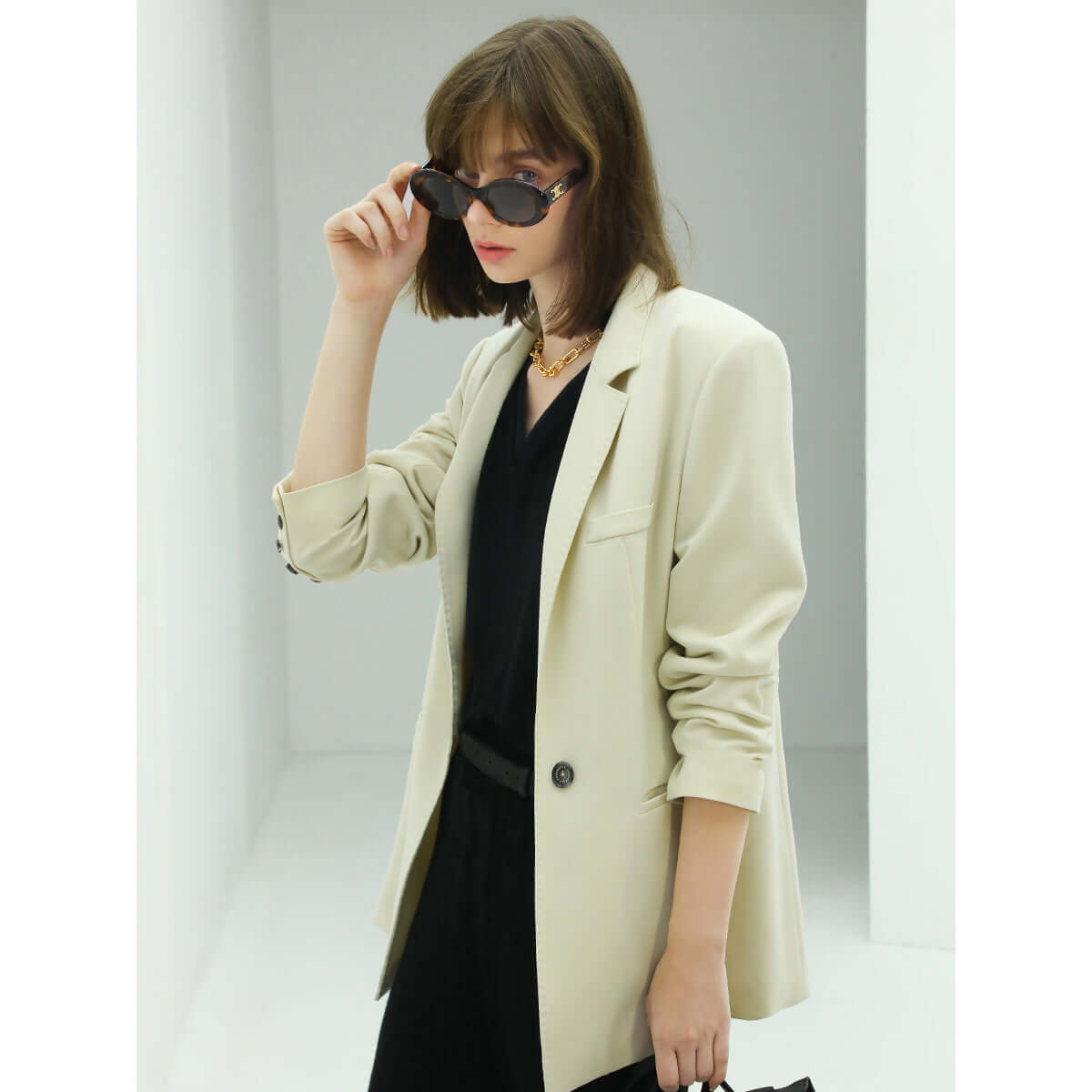 Woman wearing a beige blazer over a black dress with sunglasses, standing against a plain background.