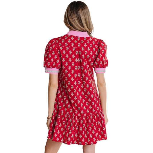 Woman wearing a red floral cotton mini dress with pink ricrac trim, V-neckline, and puff sleeves. Back view.