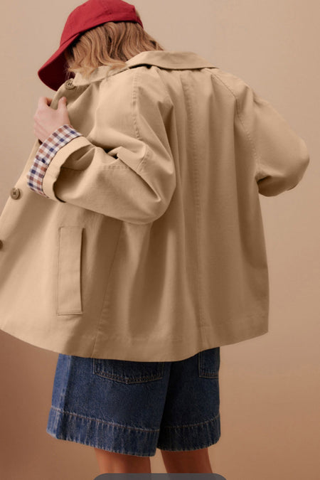 Back view of the Catherine Martin relaxed-fit beige trench coat featuring plaid-lined cuffs, styled with denim shorts and a red cap