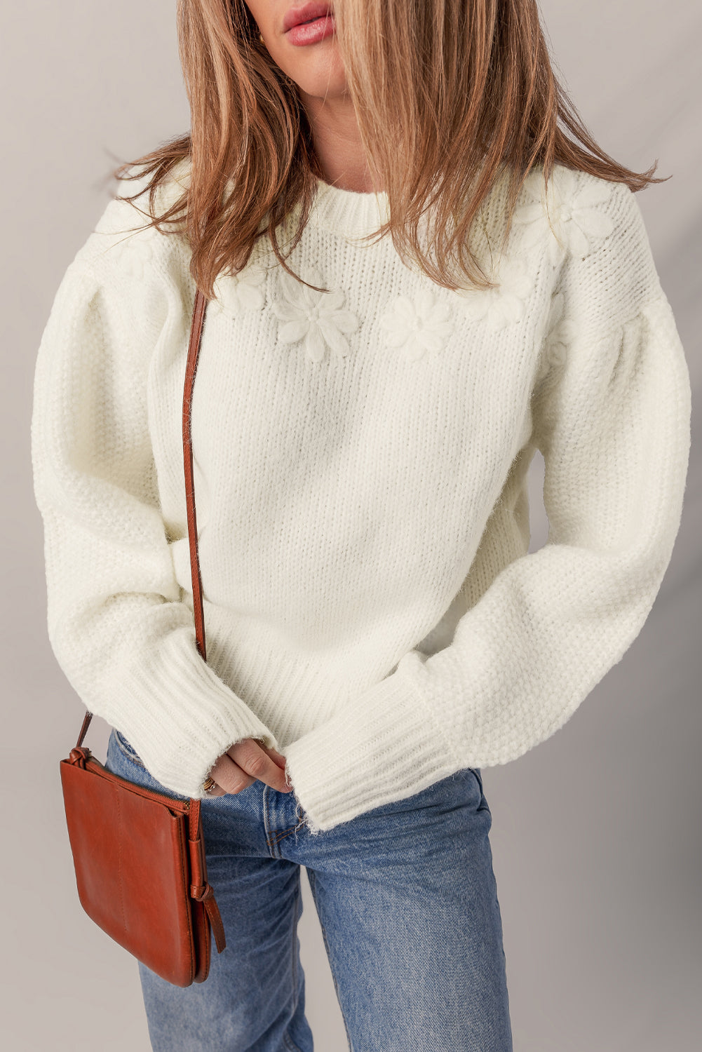 Person wearing a white sweater and blue jeans with a brown leather bag on a plain background