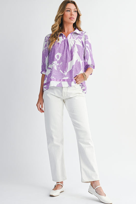 Woman wearing a purple patterned blouse and white pants on a plain background
