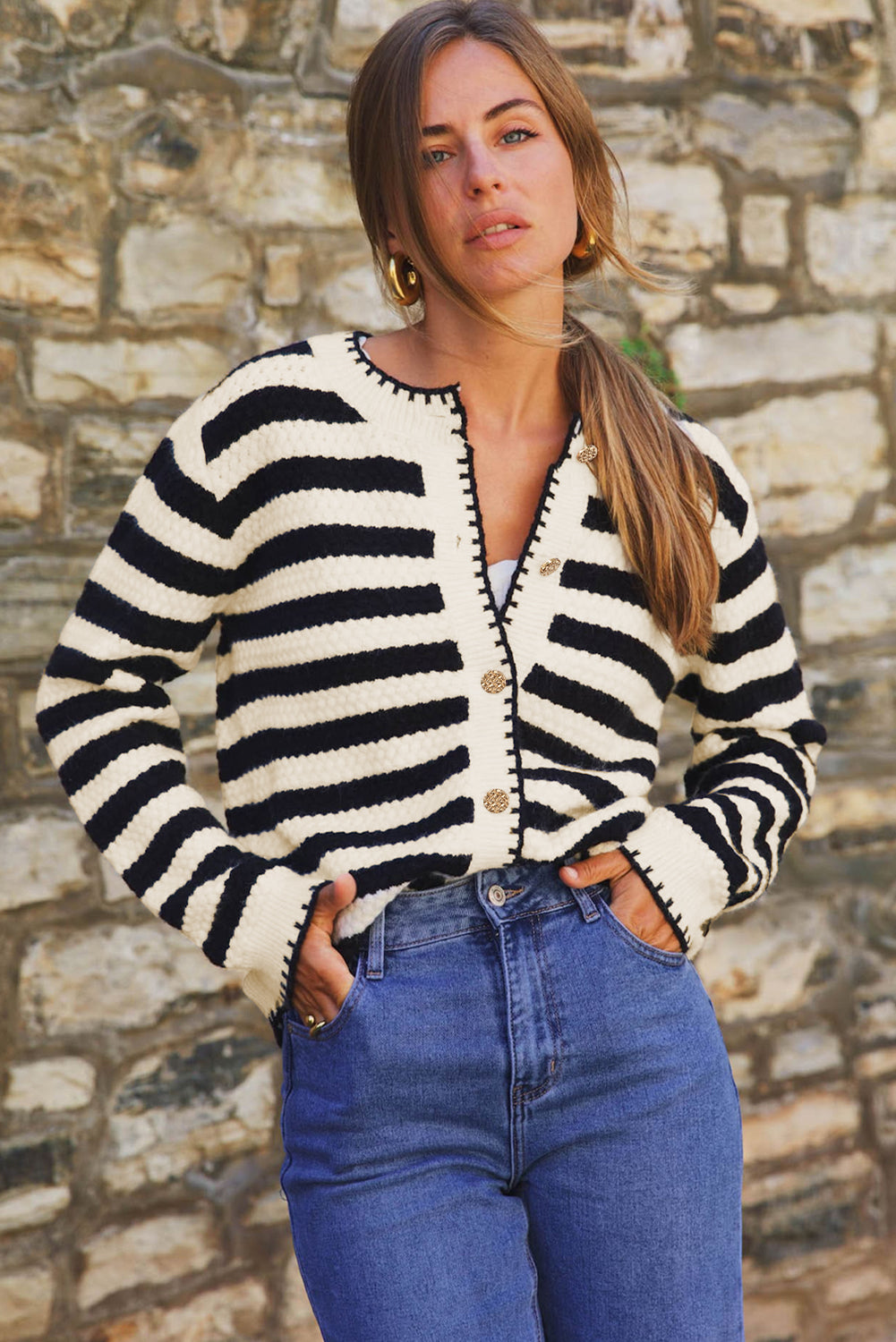 Woman wearing a black and white striped cardigan and blue jeans against a stone wall.