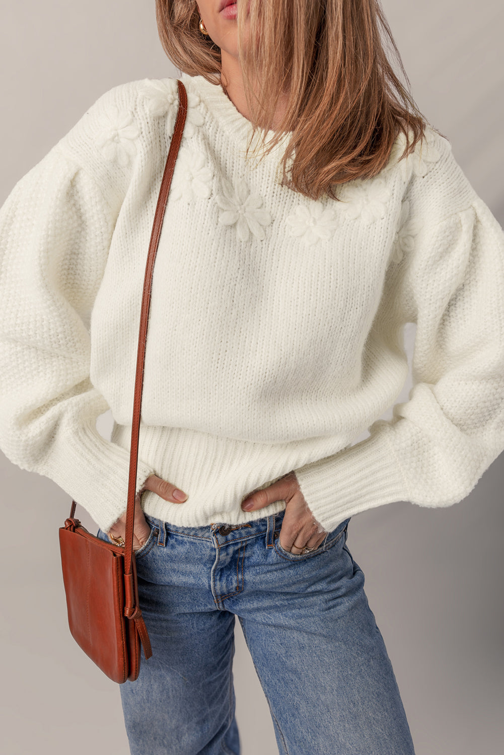 Person wearing a white sweater and blue jeans with a brown leather bag on a plain background