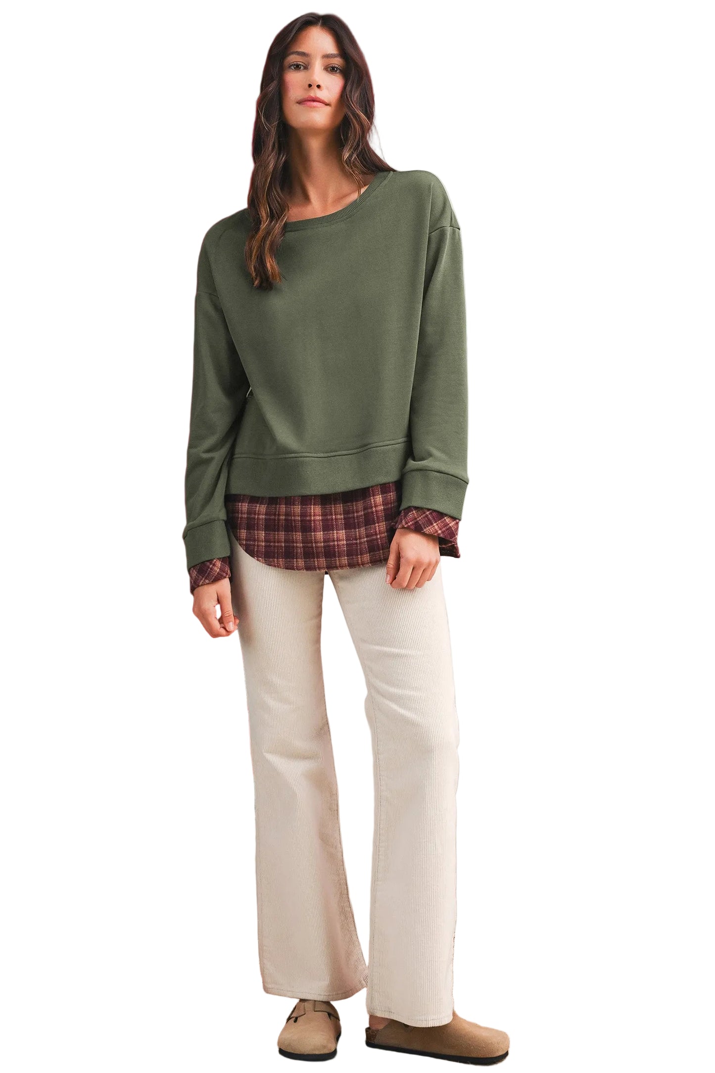 Woman in a green sweater with red brown plaid patchwork and white pants standing against a neutral white background.