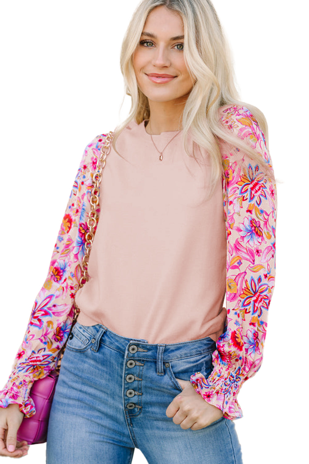 Half view of a model smiling in an oatmeal knit top with floral print long sleeves, holding a pink shoulder bag against a white background.