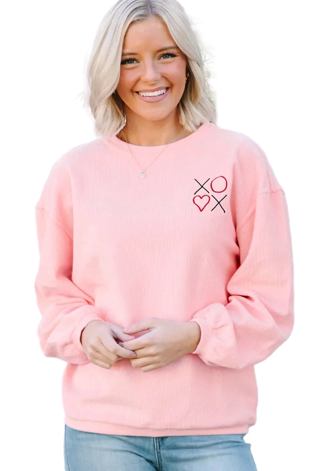 Woman wearing a pink sweatshirt with a heart and cross design, standing in front of a white lattice background.