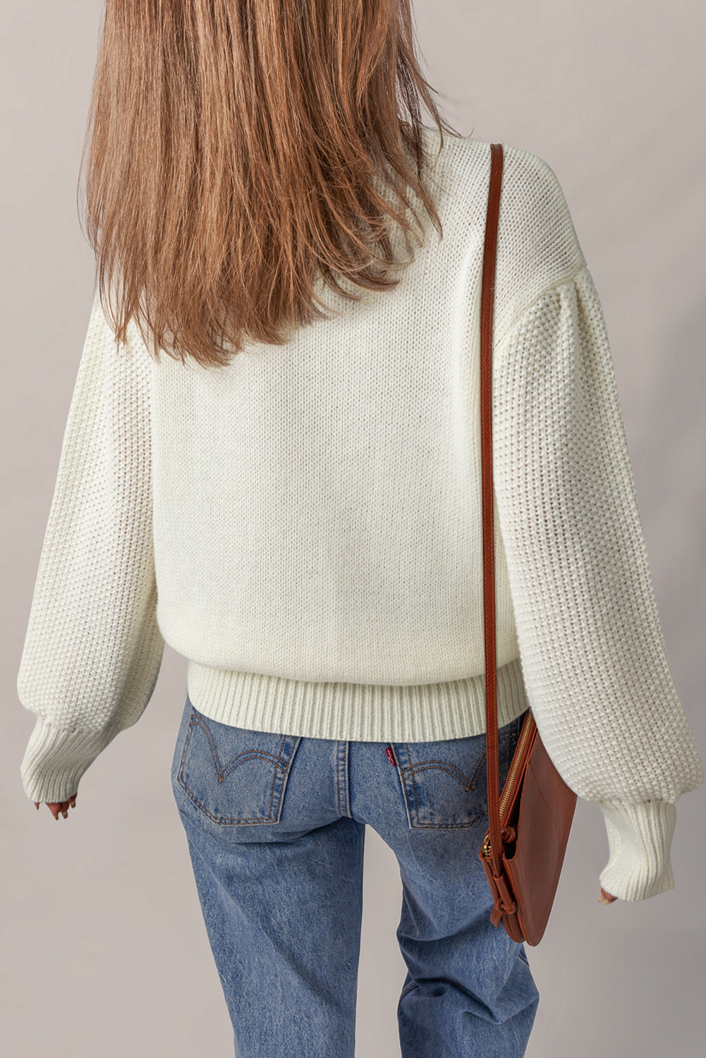Person wearing a white sweater and blue jeans with a brown leather bag on a plain background