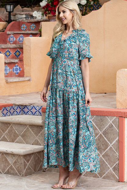 Full side view of a model standing and smiling in a paisley print tiered maxi dress with ruffle sleeves.