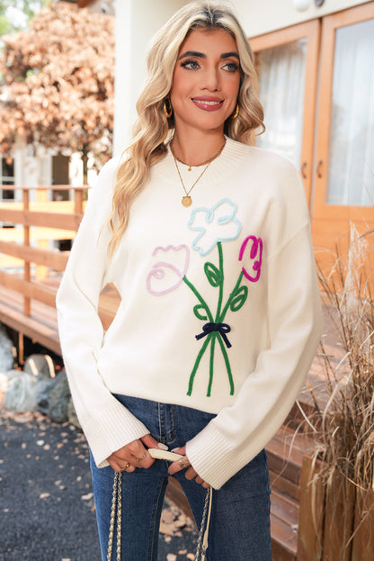 Woman wearing a cream sweater with colorful flower design outdoors.