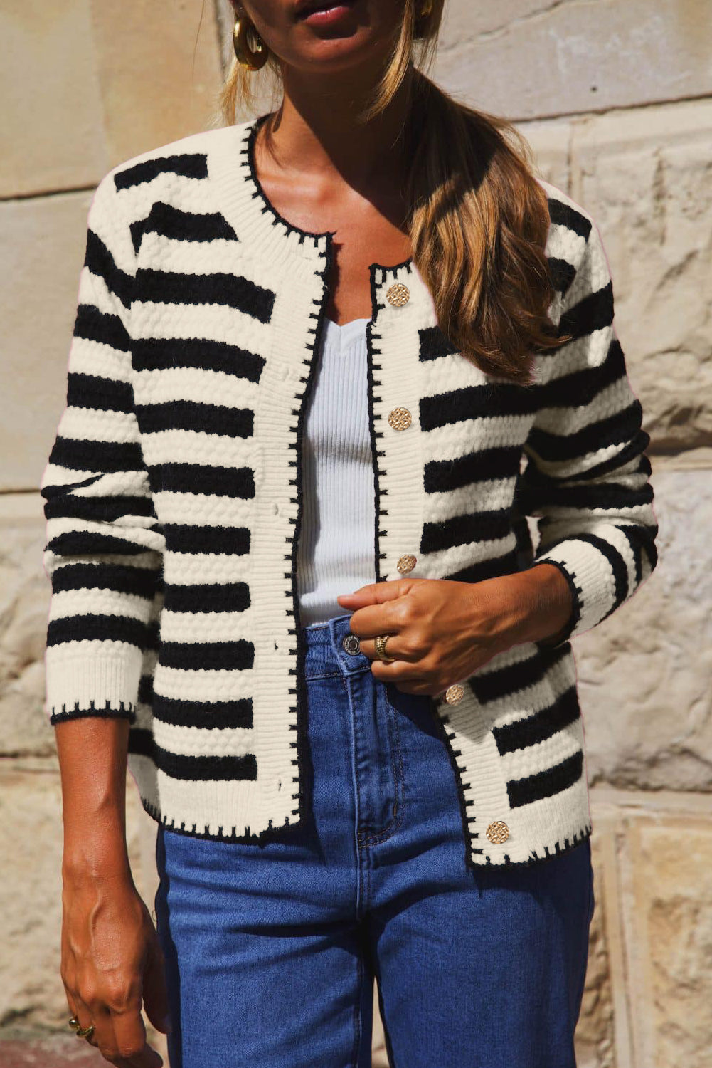 Person wearing a black and white striped cardigan with blue jeans against a stone wall background
