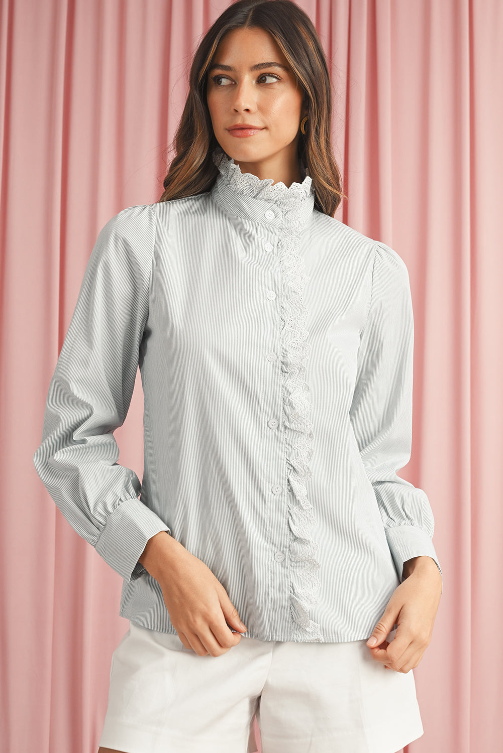 Half view of model wearing a blue striped shirt with lace trim details and long sleeves, looking to her right, set against a pink curtain background.