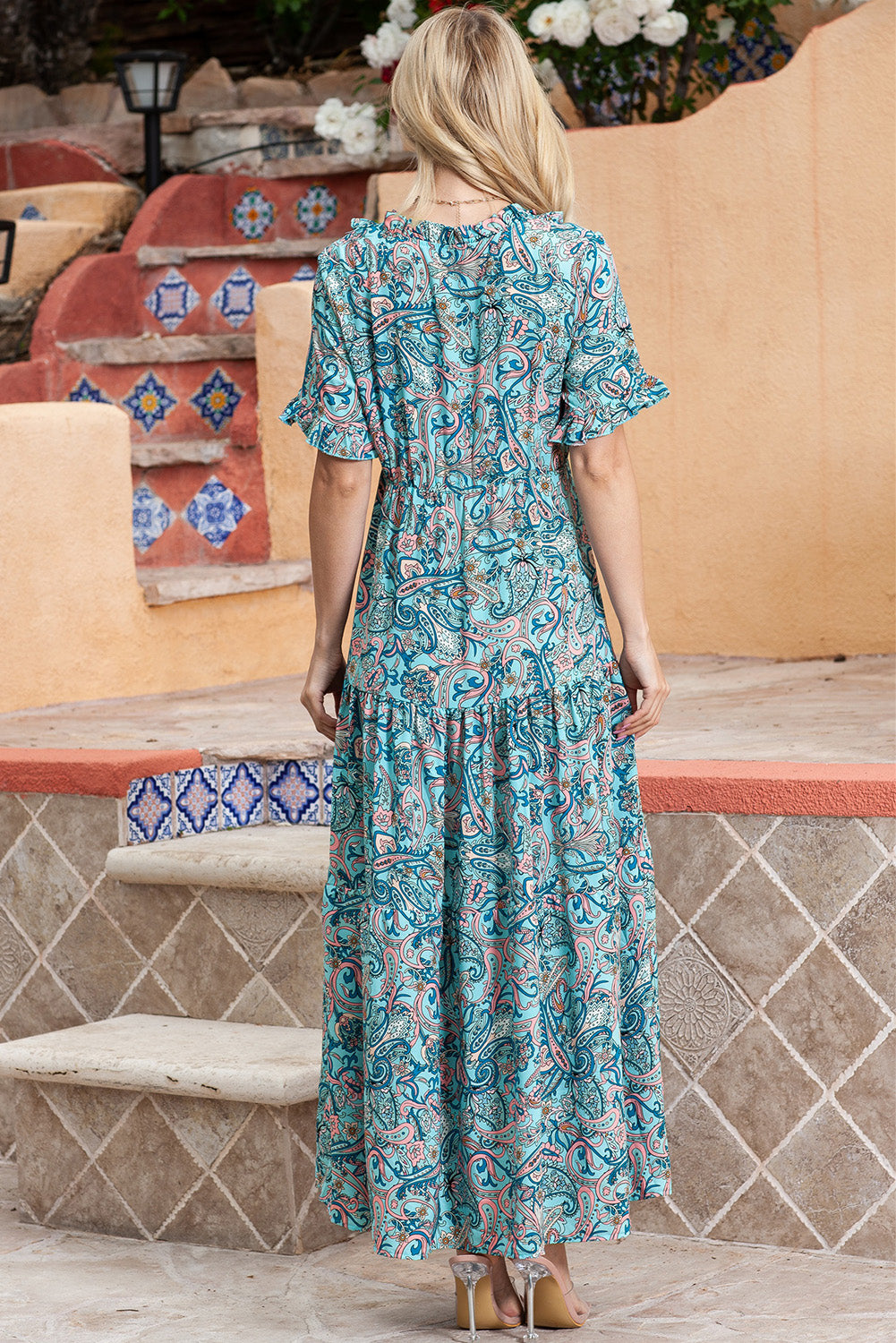 Full back view of a model standing and smiling in a paisley print tiered maxi dress with ruffle sleeves.