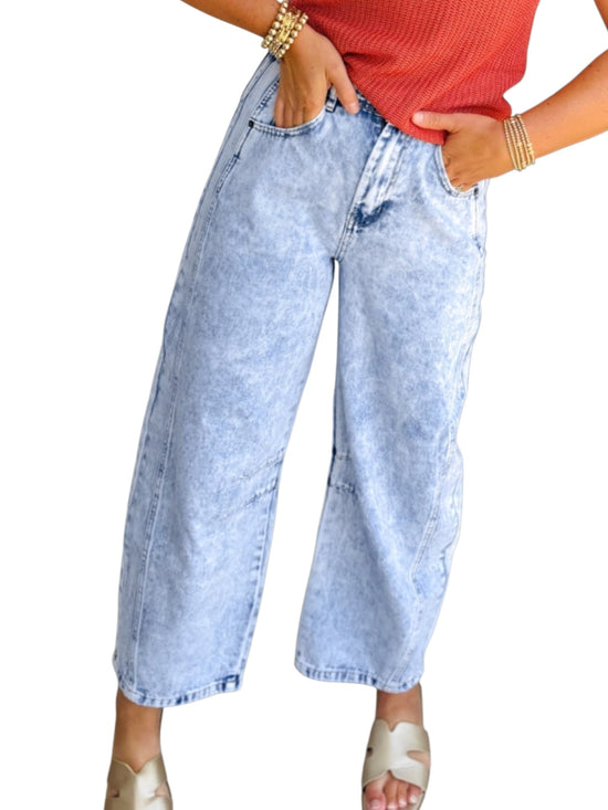 A person wearing light-wash, wide-leg cropped denim jeans with a faded blue wash, paired with a red ribbed top and beige slide sandals, shown against a plain white background.
