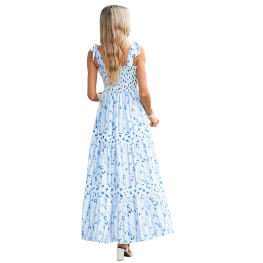 A woman wearing a blue and white striped maxi dress with a floral print, featuring a shirred bodice, tie-up ribbon shoulder straps, and a tiered flowy skirt. Back view.