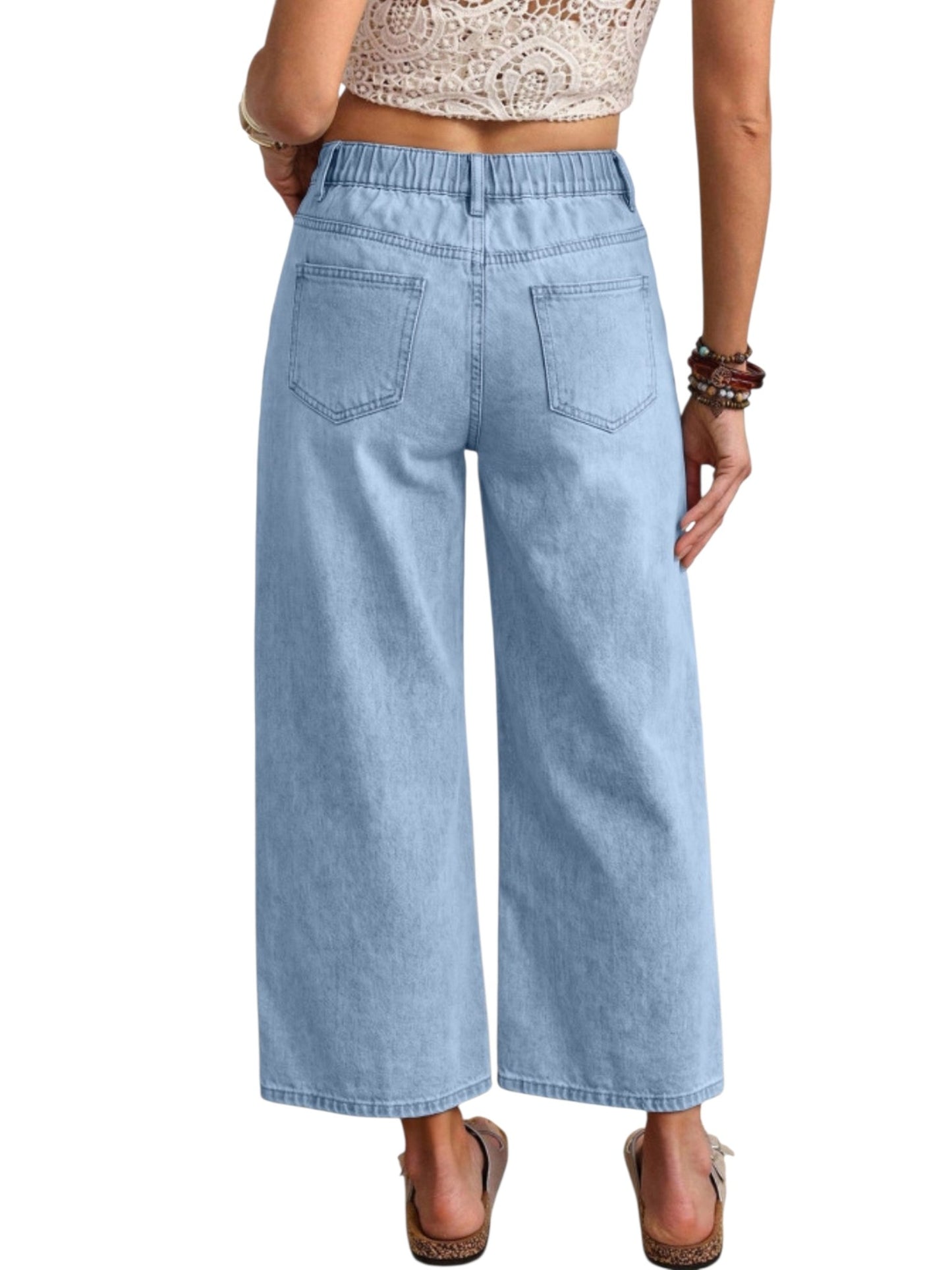 A person shown from the back wearing light-wash blue wide-leg cropped jeans, a beige lace-trim crop top, and brown wedge sandals against a plain white background.