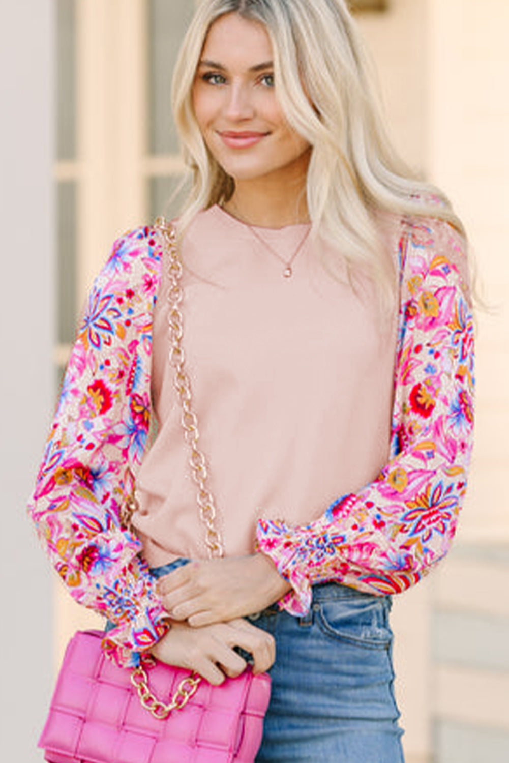 Model standing and smiling in an oatmeal knit top with floral print long sleeves, holding a pink shoulder bag.