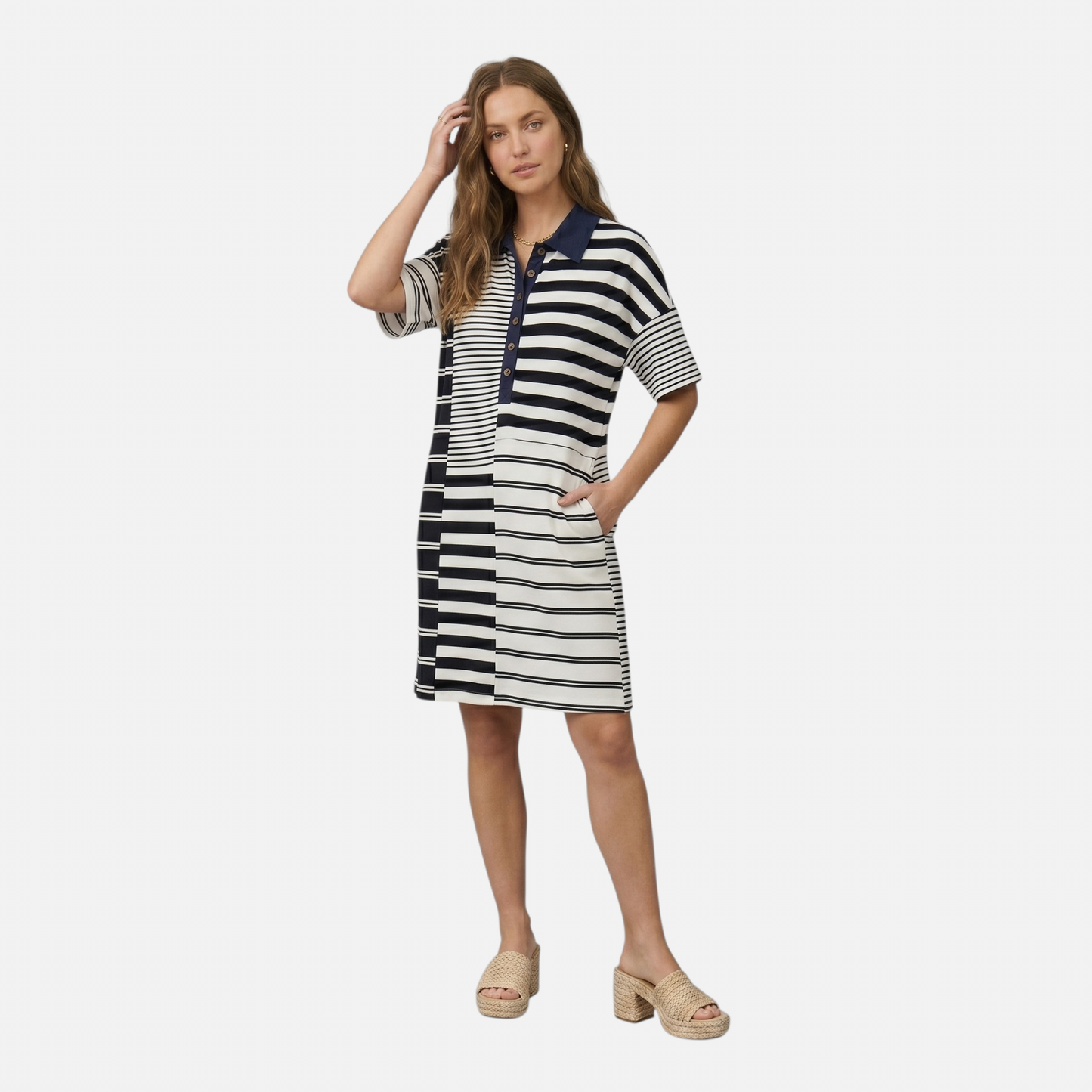 A woman posing in a navy and white striped mini dress with her hand in a side pocket. The image highlights the relaxed fit and the contrasting stripe directions across the bodice and sleeves. She is styled with natural wavy hair and woven neutral-toned sandals.