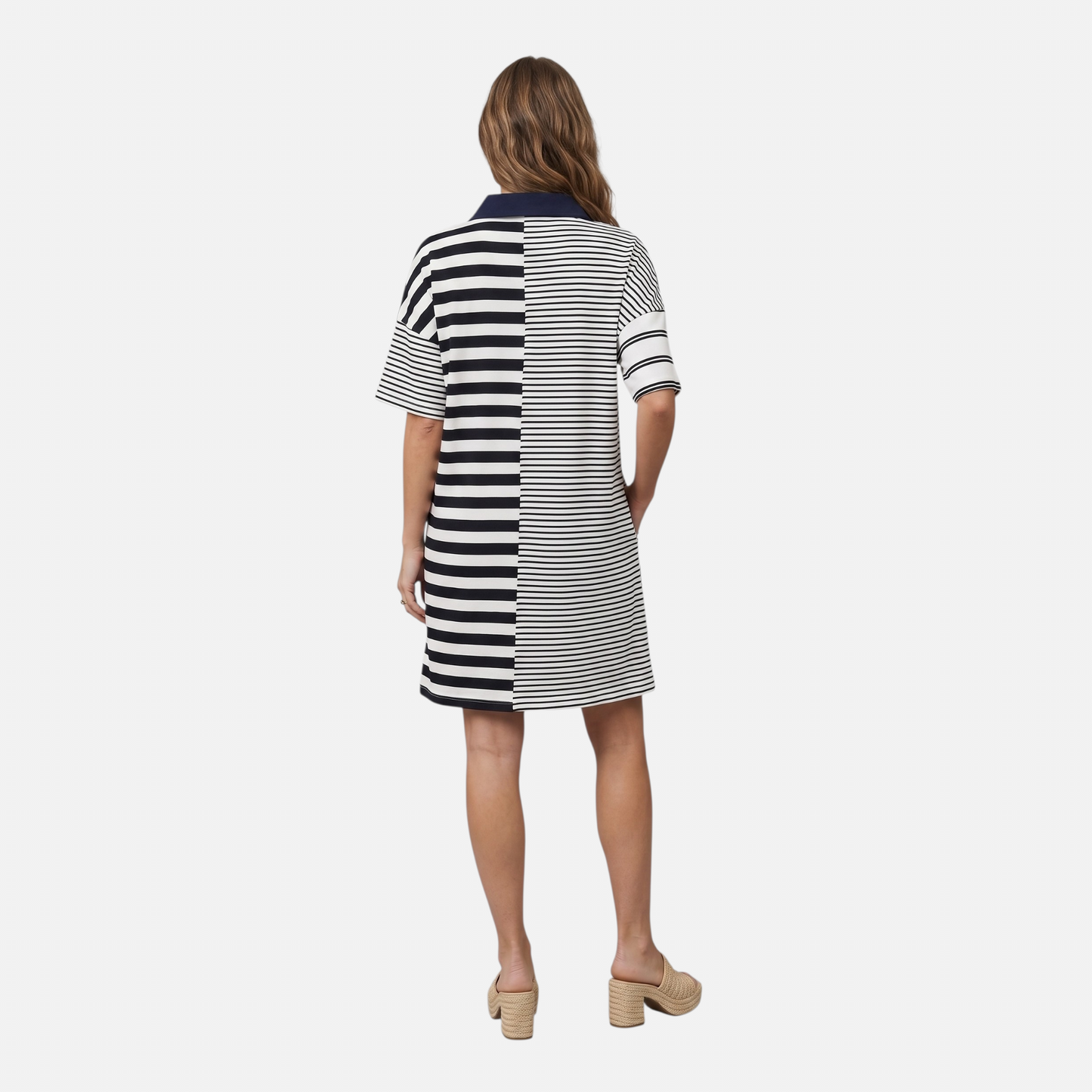 The back view of a woman in a navy and white mixed-stripe mini dress. The dress features a distinct vertical seam down the center, joining two different stripe scales for a color-block effect. She is carrying a woven straw bucket bag with a dark brown strap and wearing chunky woven heels.