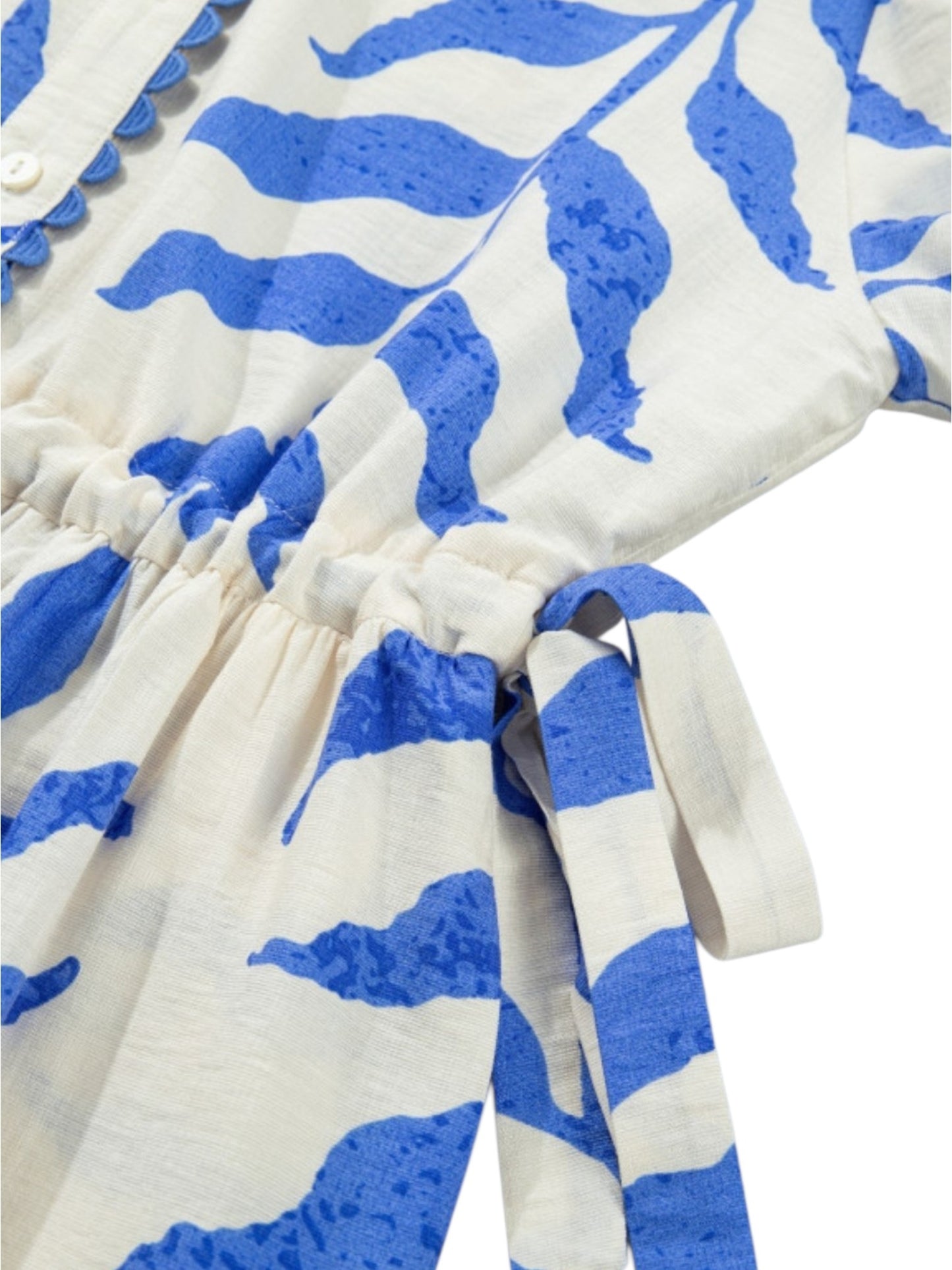 A close-up of a white cotton dress or tunic with a bold blue abstract leaf-like print, featuring a gathered waist and tie detail.