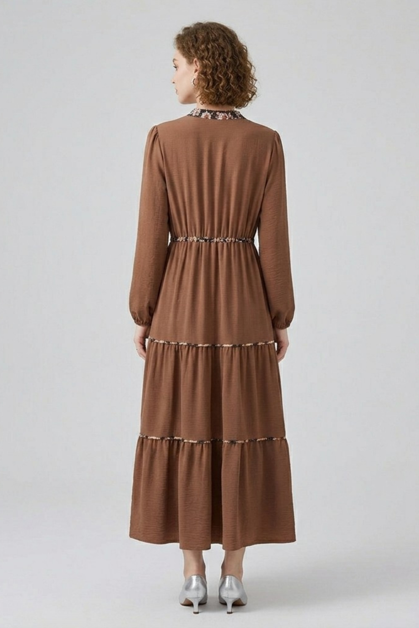 Rear view of a model in a brown tiered maxi dress, highlighting the floral print trim detailing along the collar and waistline.