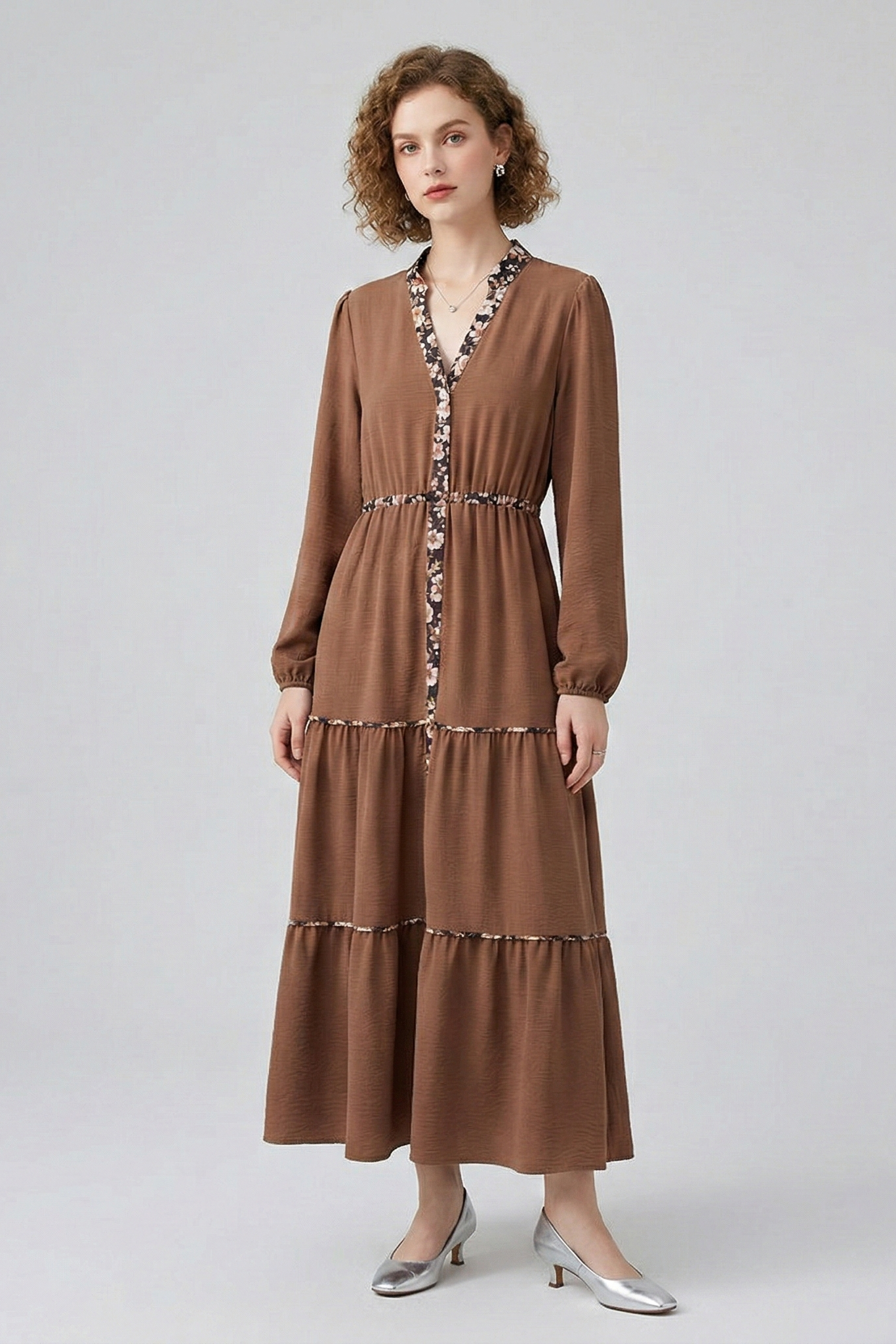 Front view of a model in a brown long-sleeved tiered maxi dress, featuring delicate floral print detailing along the V-neck, placket, and tiered seams.