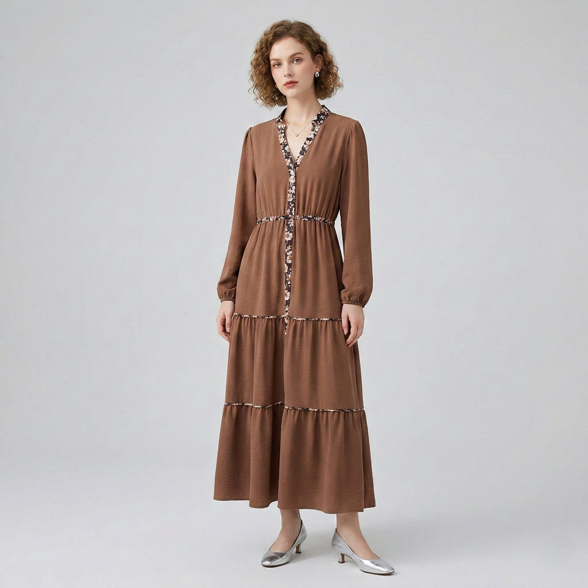 Front view of a model in a brown long-sleeved tiered maxi dress, featuring delicate floral print detailing along the V-neck, placket, and tiered seams.