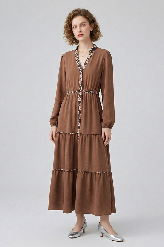 Front view of a model in a brown long-sleeved tiered maxi dress, featuring delicate floral print detailing along the V-neck, placket, and tiered seams.