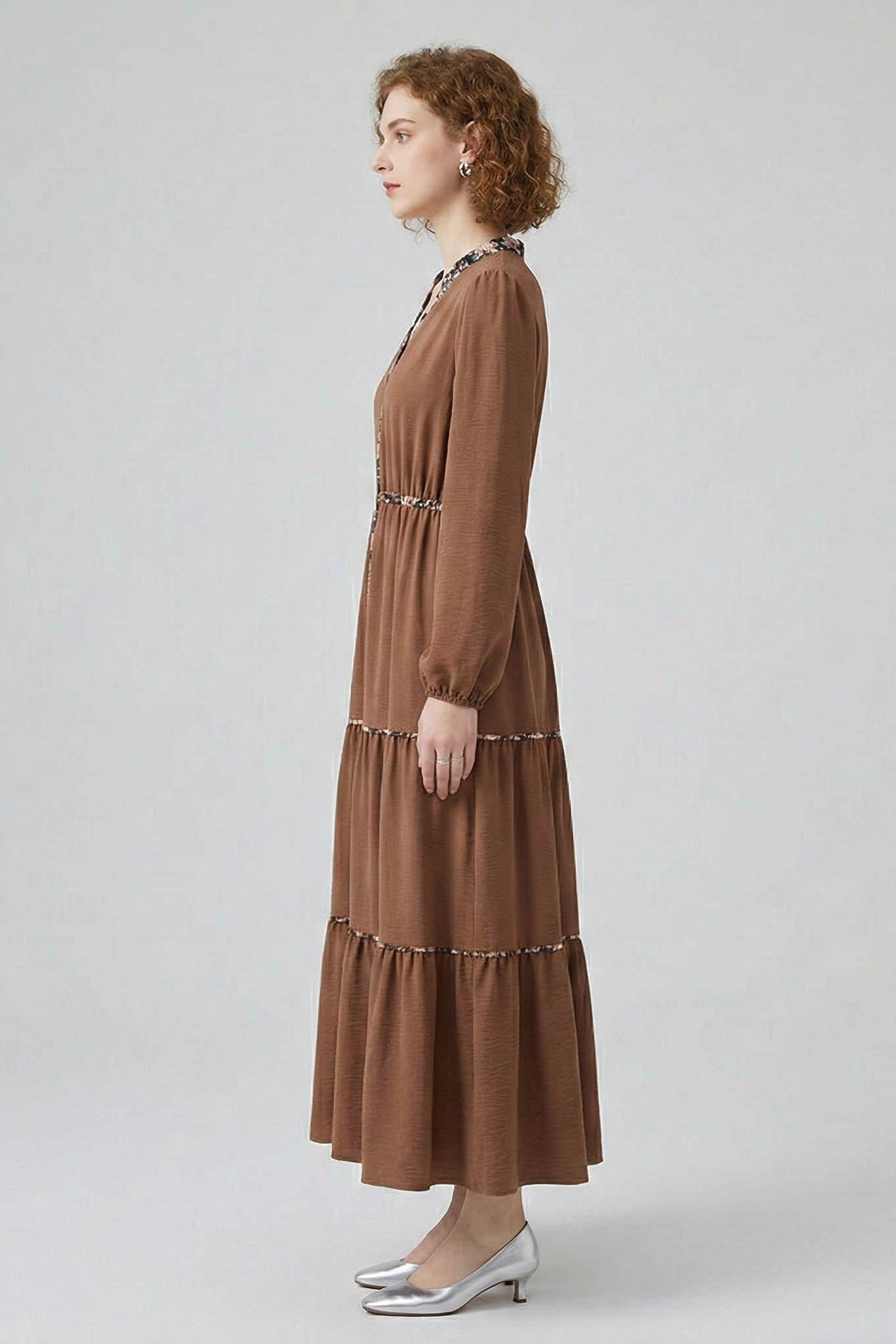 Side profile view of a model wearing a brown tiered maxi dress, showcasing the flowy, full-length silhouette and long-sleeved design with subtle floral trim accents.