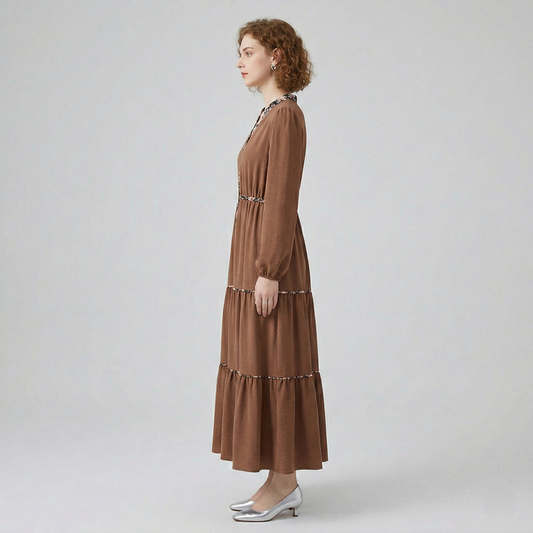 Side profile view of a model wearing a brown tiered maxi dress, showcasing the flowy, full-length silhouette and long-sleeved design with subtle floral trim accents.