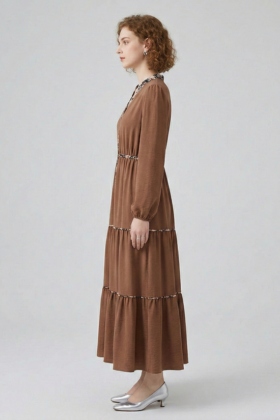 Side profile view of a model wearing a brown tiered maxi dress, showcasing the flowy, full-length silhouette and long-sleeved design with subtle floral trim accents.