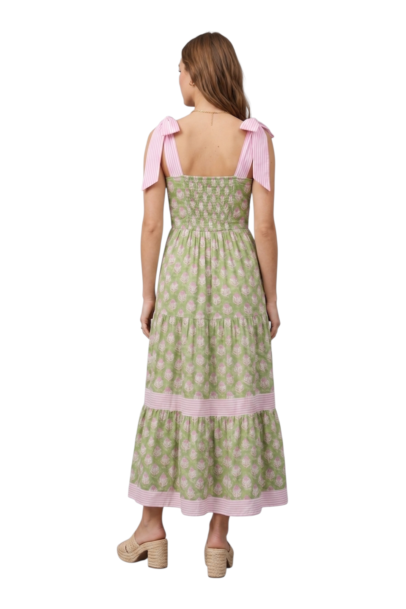 Woman wearing a green and pink botanical block print tiered maxi dress with pink striped tie-knot shoulder straps and smocked back bodice, paired with tan woven platform mule heels, back view