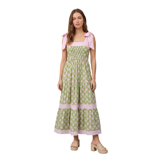Woman wearing a green and pink botanical block print tiered maxi dress with pink striped tie-knot shoulder straps, smocked bodice, and pink striped hem trim, paired with tan woven platform mule heels, front view