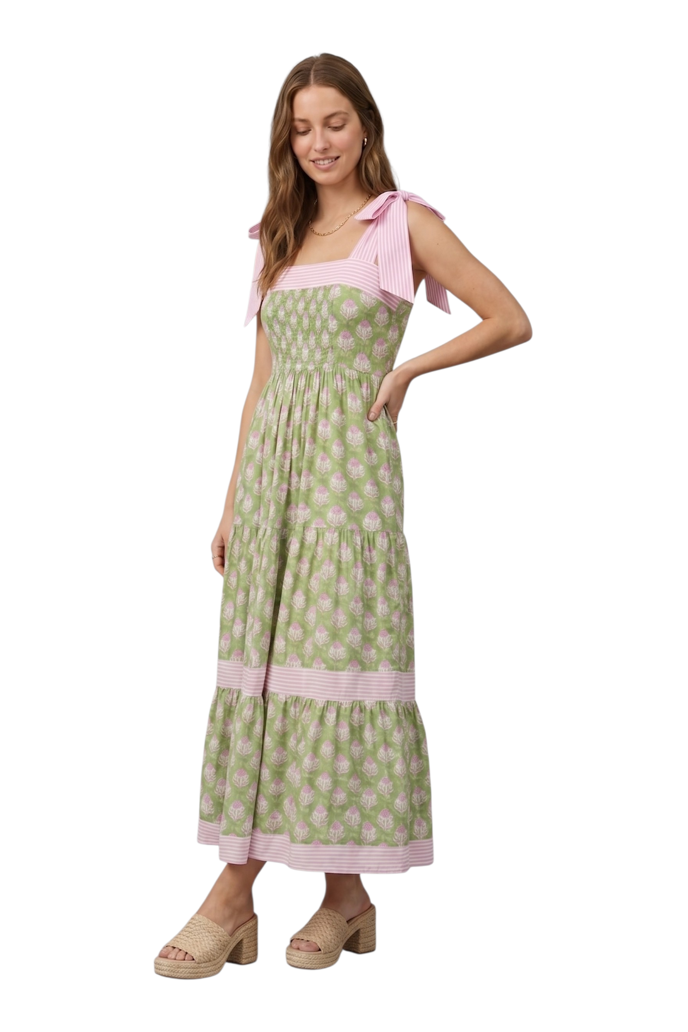 Woman wearing a green and pink botanical block print tiered maxi dress with pink striped tie-knot shoulder straps and smocked bodice, hand in pocket, looking down, three-quarter view