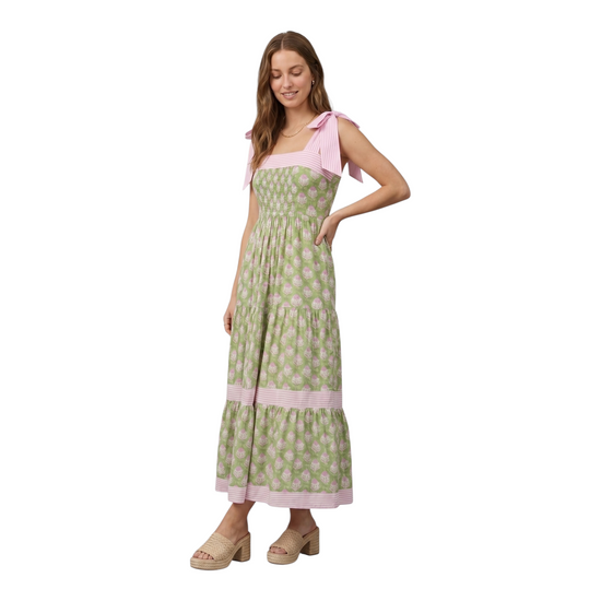 Woman wearing a green and pink botanical block print tiered maxi dress with pink striped tie-knot shoulder straps and smocked bodice, hand in pocket, looking down, three-quarter view
