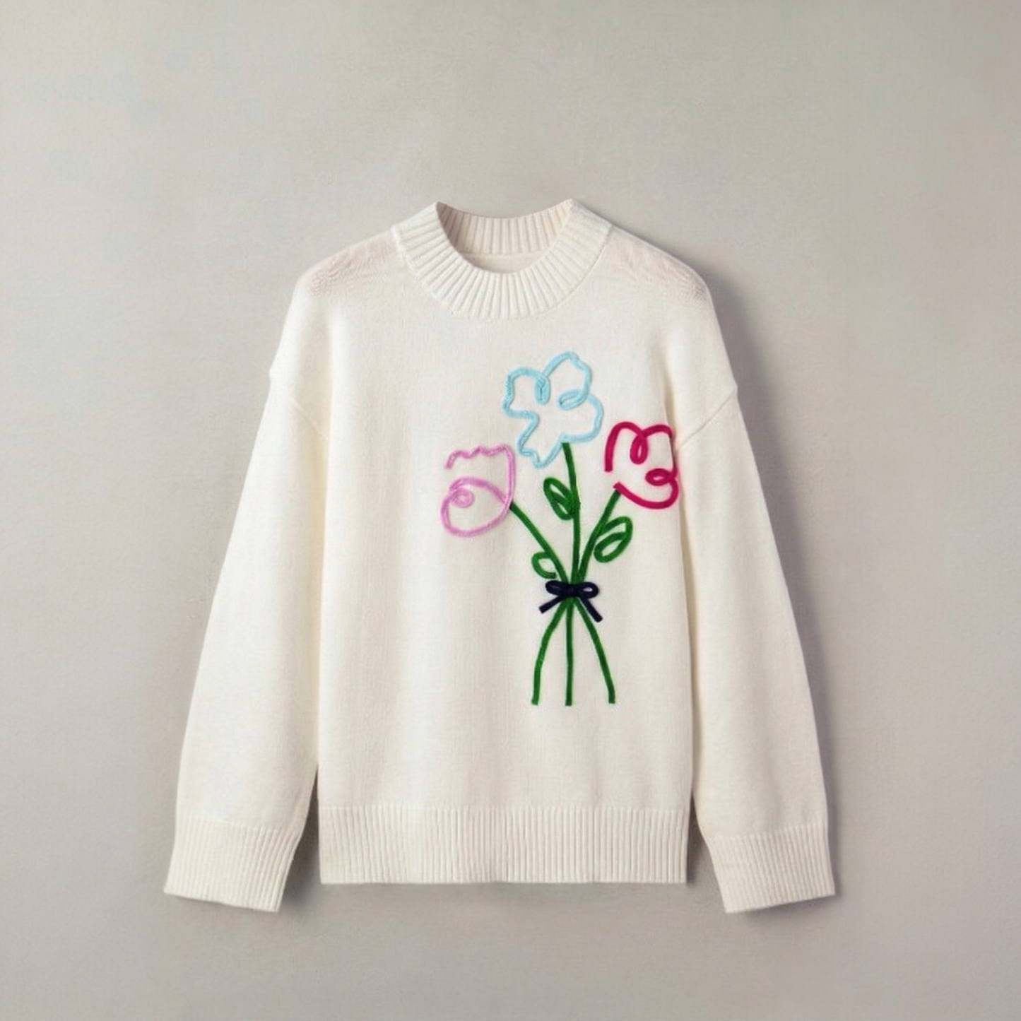 Front view of a white knit sweater featuring a colourful embroidered flower bouquet on the chest, displayed on a wooden hanger.