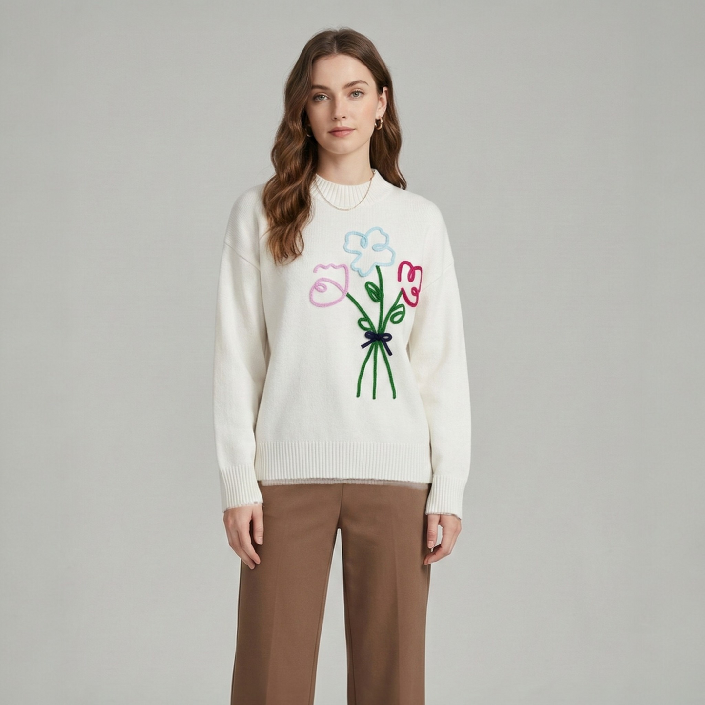 A model wearing a white drop-sleeve sweater with a colourful flower bouquet embroidery on the chest, styled with brown trousers.