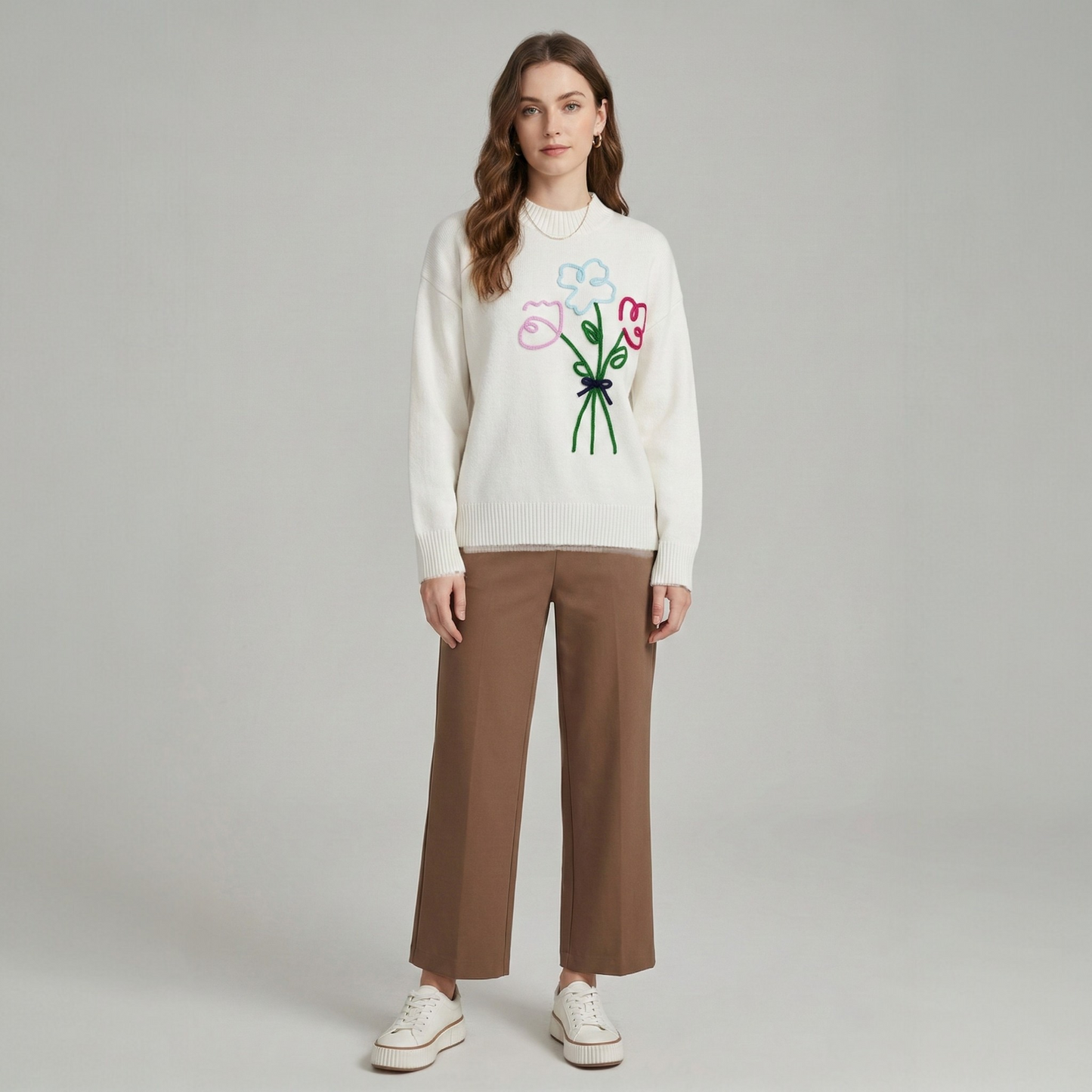 Full-length front view of a model wearing a white knit sweater with bouquet embroidery, styled with brown trousers and white sneakers.