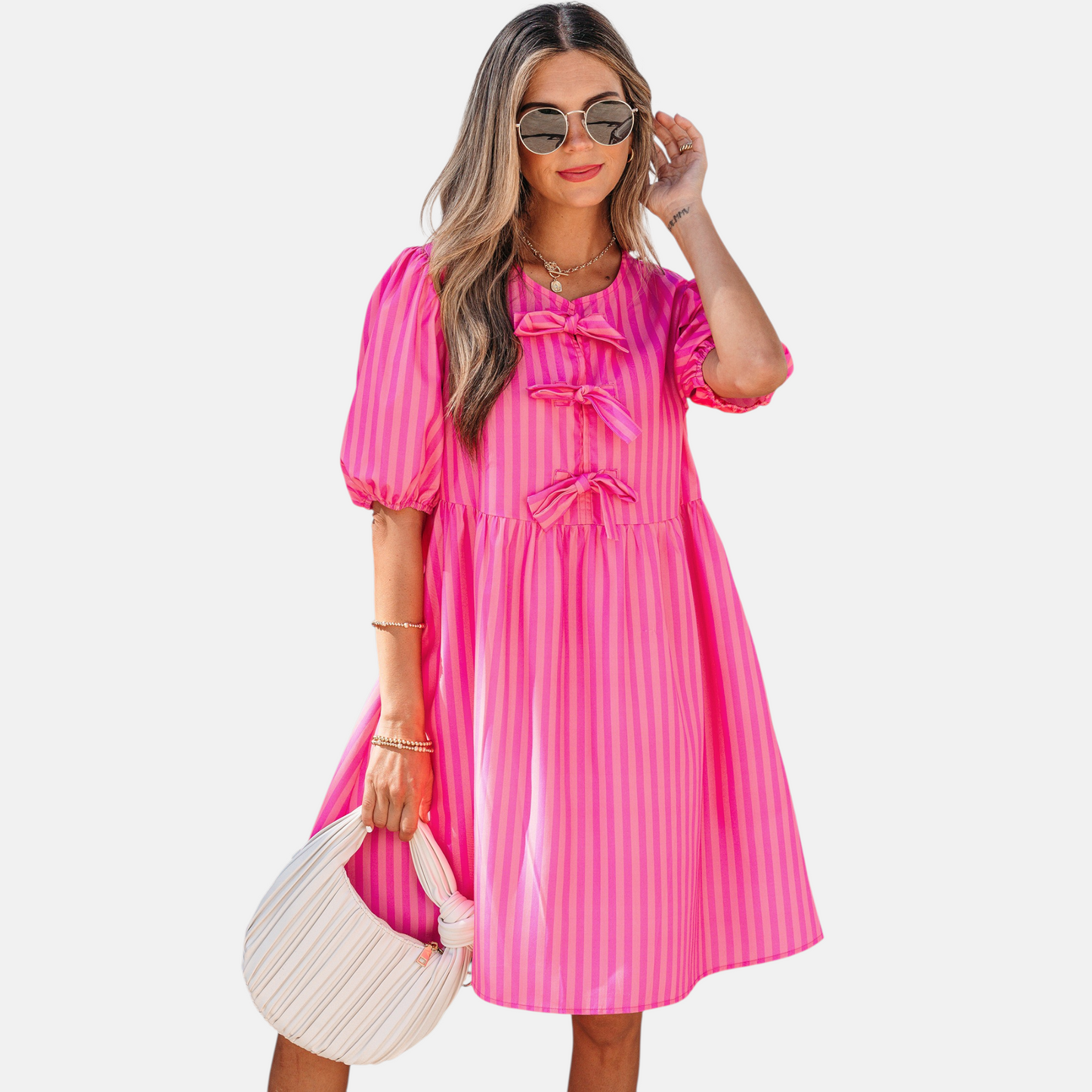 A model posing in a vibrant pink striped mini dress featuring puff sleeves, three decorative front bows, and functional side pockets, styled with a pleated handbag.