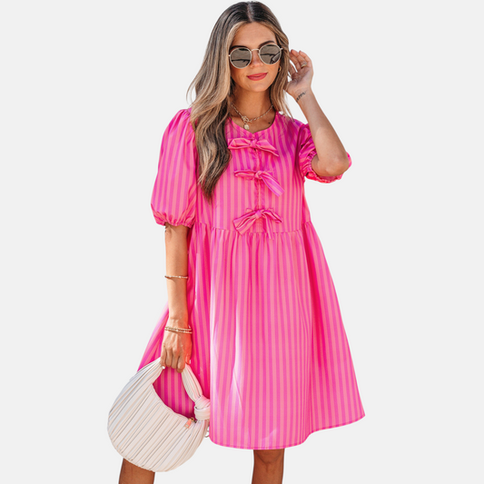 A model posing in a vibrant pink striped mini dress featuring puff sleeves, three decorative front bows, and functional side pockets, styled with a pleated handbag.